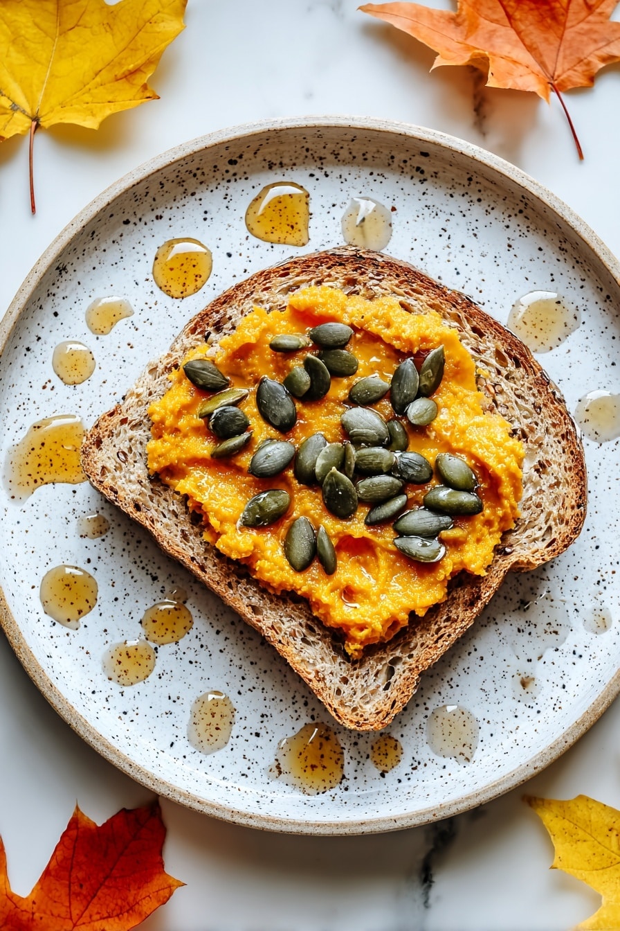 A single slice of toasted multigrain bread with a rough texture sits at the center of a white speckled plate. On top of the bread is a thick layer of bright orange pumpkin spread with a slightly chunky texture. Scattered evenly across the pumpkin spread are dark green pumpkin seeds that add a glossy contrast. Around the bread on the plate are small drops of honey, creating a shiny amber pattern. The plate rests on a white marbled surface with scattered bright orange and yellow autumn leaves, adding a seasonal touch. photo taken with an iphone --ar 2:3 --v 7 - Spiced Pumpkin Cream Cheese Toast, pumpkin toast recipe, fall breakfast ideas, cozy pumpkin toast, easy autumn breakfast