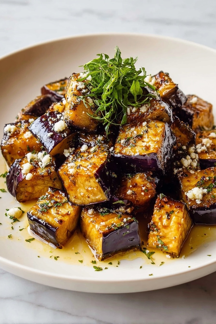 The dish shows about two layers of cubed eggplant pieces in a white plate. The eggplant cubes have a dark purple skin on the edges and soft golden-brown flesh on the inside with a shiny glaze of oil. There are small white bits sprinkled on the cubes that look like coarse salt or cheese, along with green herb flakes. A small bunch of fresh green herbs is placed in the center of the pile on the top layer. The plate sits on a white marbled surface. photo taken with an iphone --ar 2:3 --v 7 - Crispy Garlic Roasted Eggplant, roasted eggplant side dish, garlic eggplant recipe, crunchy eggplant appetizer, healthy roasted vegetable dish