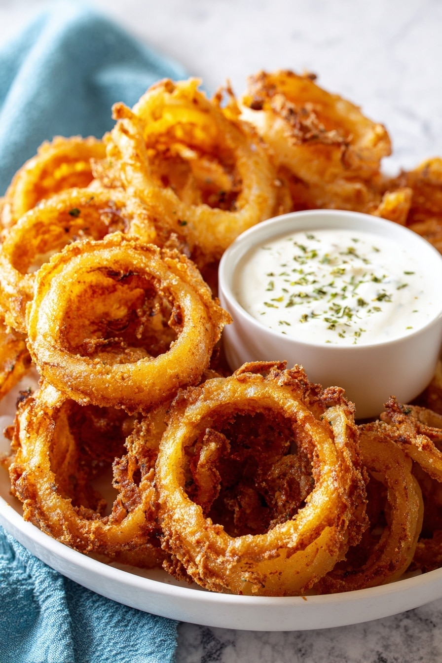A white bowl is filled with many golden brown, crispy fried onion rings that have a bubbly texture along the edges and a slightly oily shine. They are layered unevenly, showing natural onion rings with a light crisp dusting. In the center of the bowl sits a smaller white bowl filled with white creamy sauce topped with small green herb pieces scattered on top. The background is a white marbled surface and there is a soft blue cloth visible to the side. photo taken with an iphone --ar 2:3 --v 7 - Crispy Onion Ring Chips, onion chip snack, crunchy onion chips, homemade onion rings, easy onion ring recipe