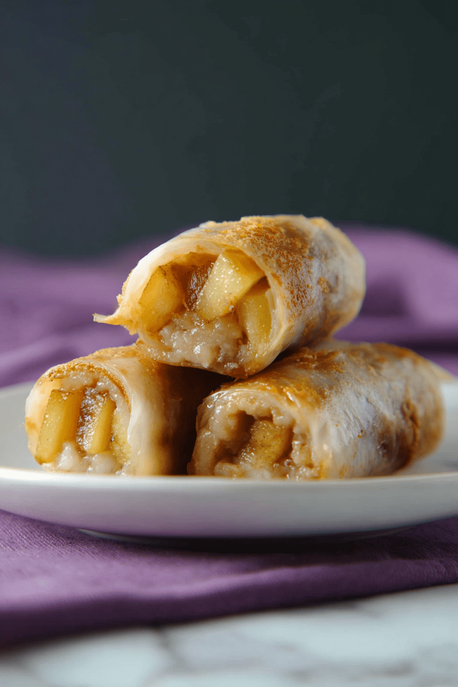 Four pieces of rolled dessert filled with a soft, cooked filling that looks like sliced yellow fruit mixed with spices. Each piece is light brown and slightly shiny with a thin outer layer that looks soft and slightly sticky. The pieces are stacked in two layers on a white plate, with the background showing a white marbled texture under the plate and a dark blurred backdrop. The plate itself rests on a purple cloth. photo taken with an iphone --ar 2:3 --v 7 - Baked Apple Rice Paper Rolls, baked apple spring rolls, apple dessert rolls, crispy baked fruit rolls, cinnamon apple snacks